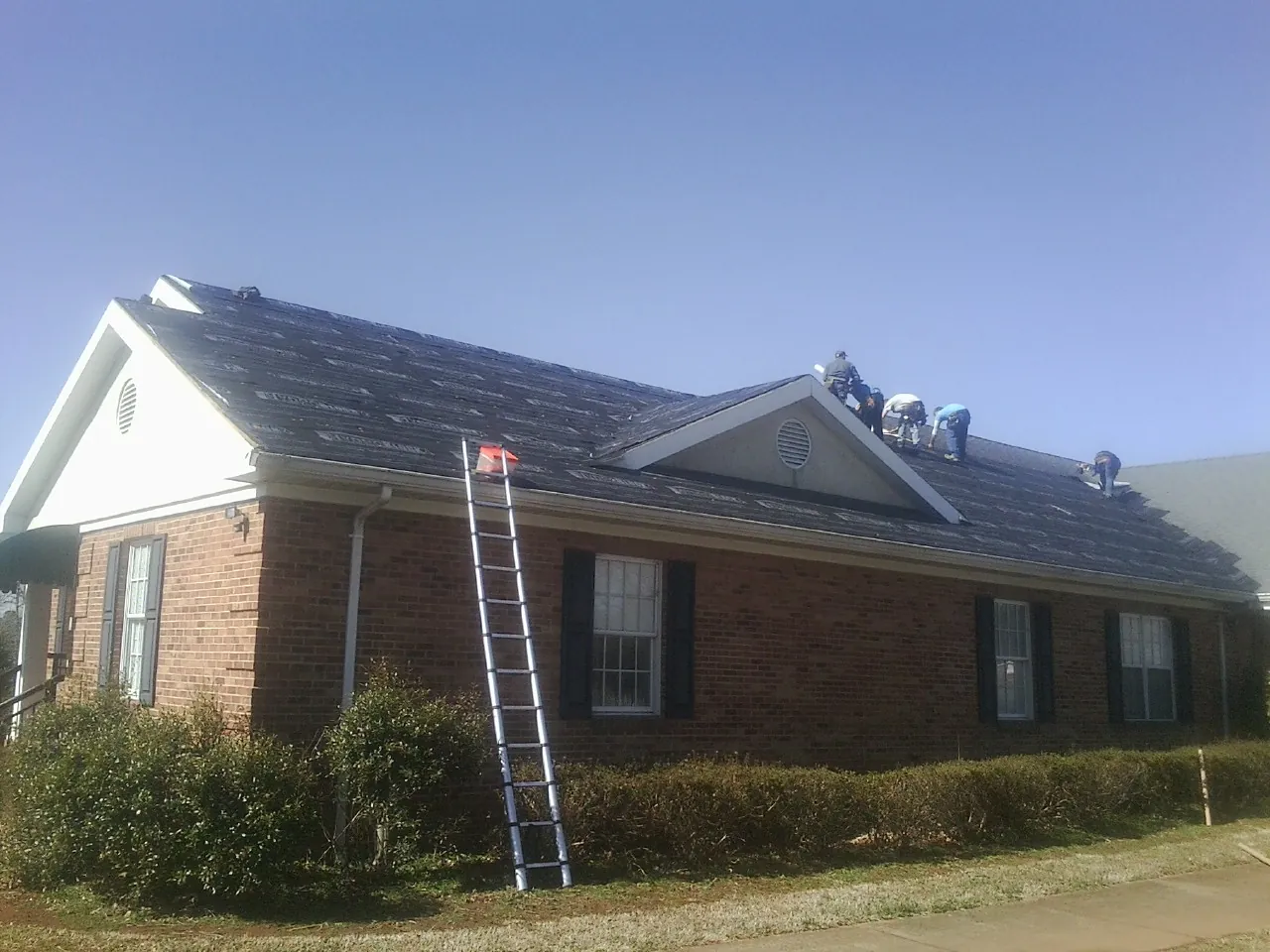 Crew tearing shingles off a brick ranch roof with a ladder set — Emergency Roof Repair work by our crew in Wirtz