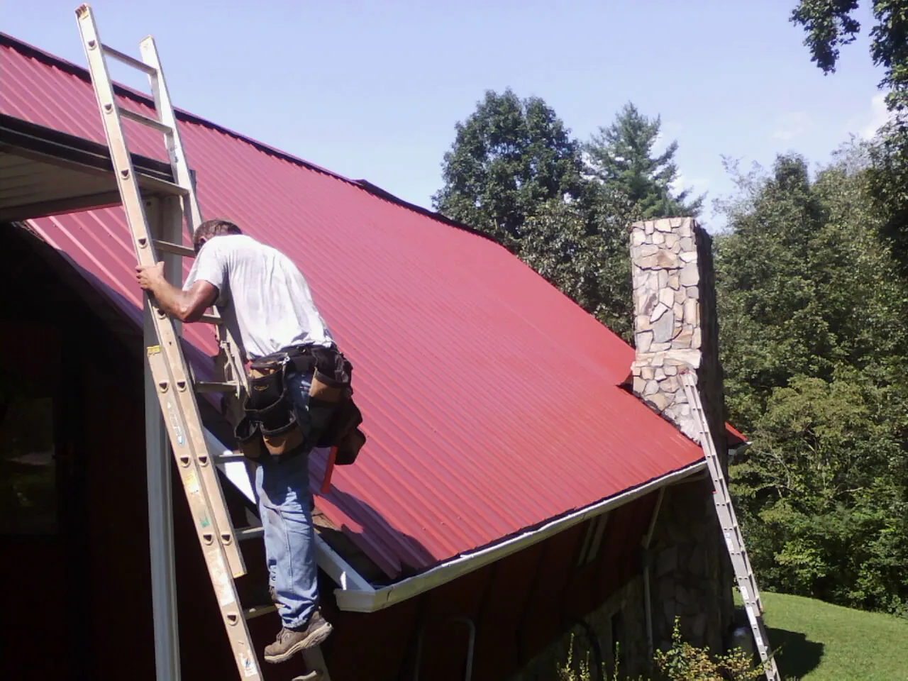 Roofer on a ladder beside a red metal roof and stone chimney — Soffit & Fascia Repair by Roofing Brothers in Wirtz